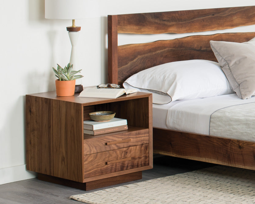 Wooden nightstand next to a bed with white bedding and wooden headboard.