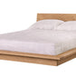 A modern low-profile platform bed with a light Oak wood frame and headboard, dressed in white bedding and two white pillows, shown against a plain white background.