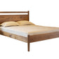 A modern Walnut wooden bed frame with a headboard and footboard, styled with a mattress and two pillows, shown on a plain white background.