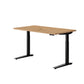 An adjustable standing desk with a light Oak wood grain top and black metal legs, shown on a solid white background.